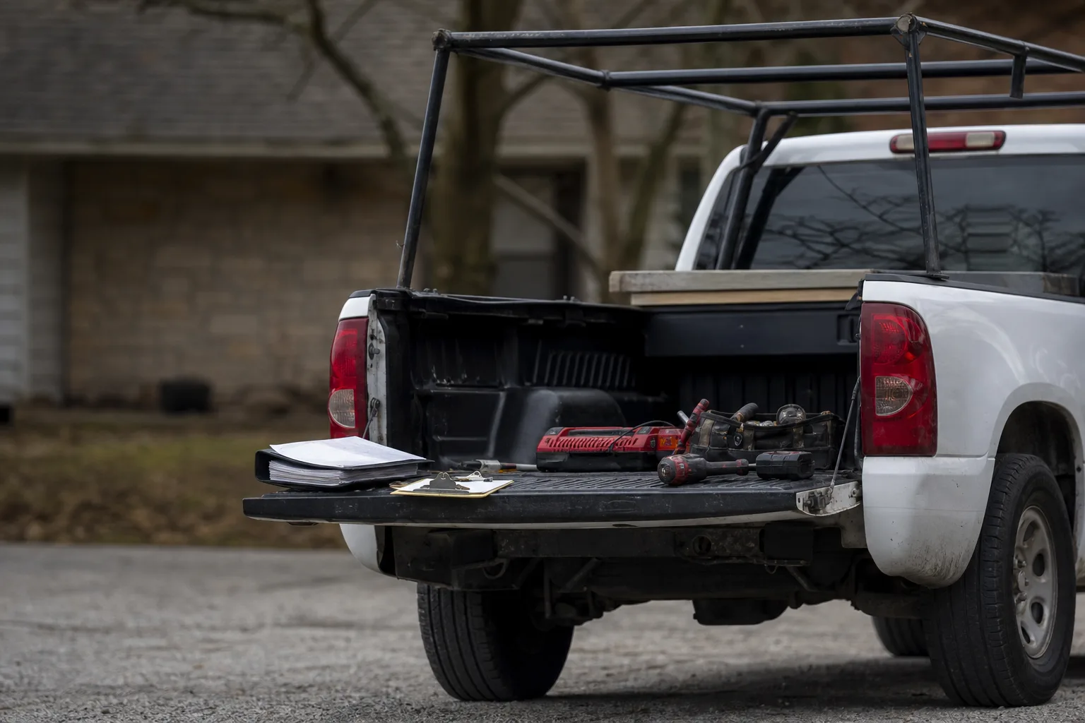 A work truck parked in a residential driveway with the tailgate down, a ladder rack overhead, and a folder of paperwork and a clipboard resting on the tailga...