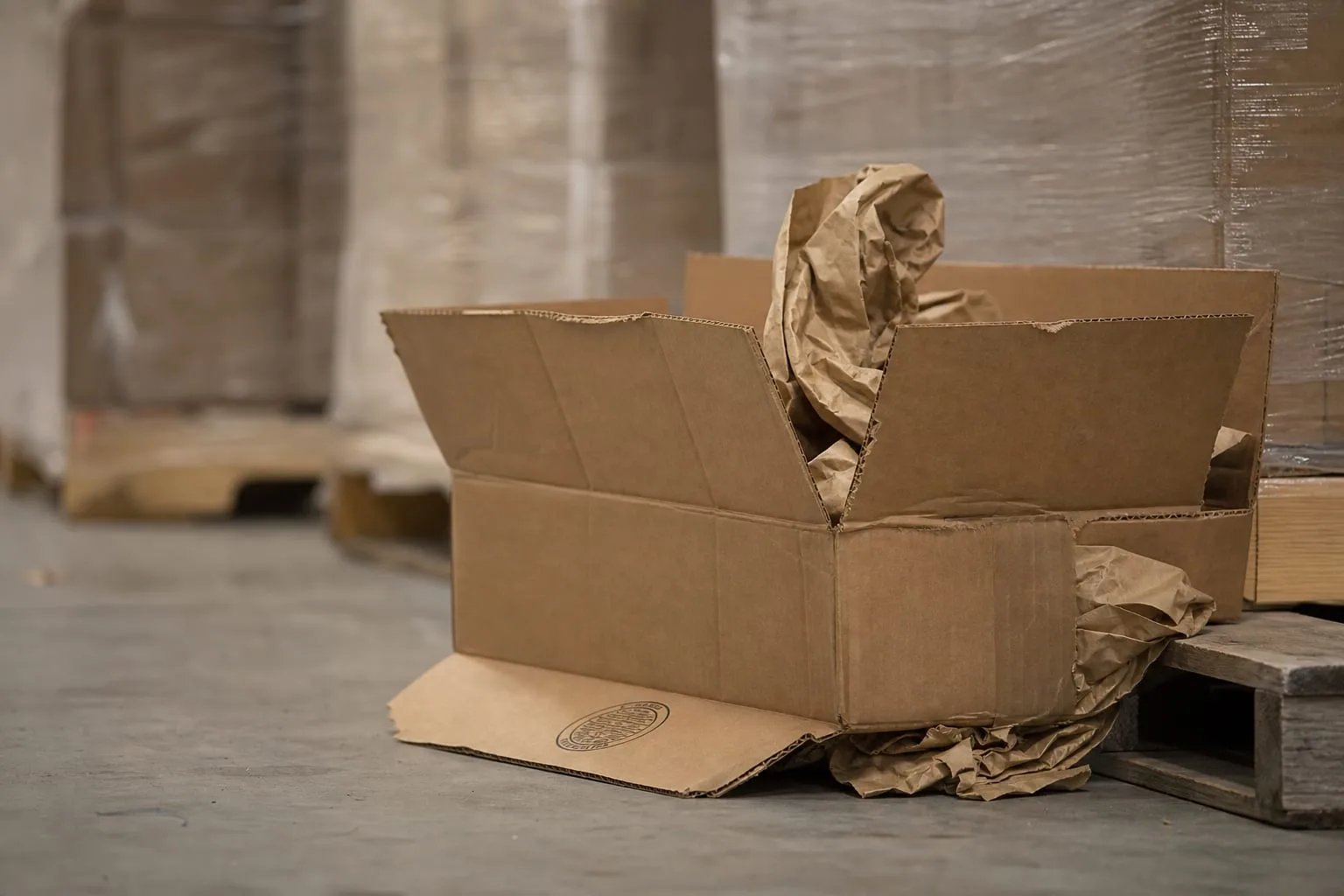 A split double-wall corrugated shipping box on a warehouse floor beside a pallet, with the box certificate stamp visible on the bottom flap and packing paper...