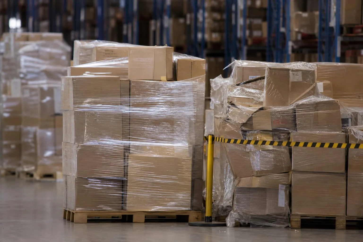 A palletized shipment of retail cartons in a contract warehouse aisle, with a quarantine area of dented and shrink-wrapped returns visible nearby