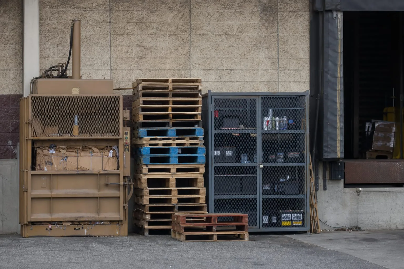 The rear yard of a distribution warehouse showing a cardboard baler, stacked wooden pallets sorted by grade, a locked cage of batteries and aerosols, and an...