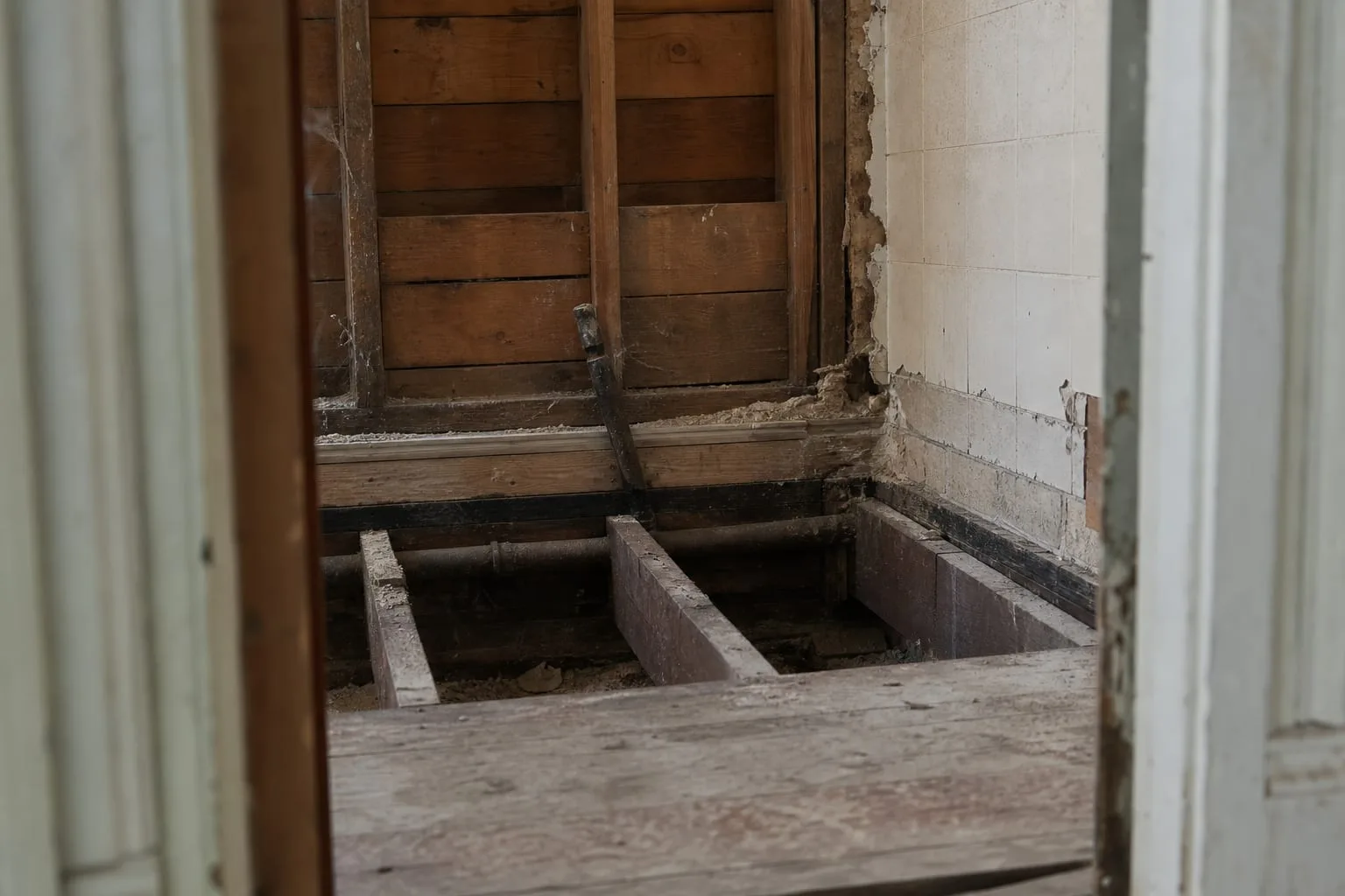A bathroom stripped to framing around a removed cast iron tub, showing exposed floor joists, a cut section of subfloor, and the edge of old tile still on the...